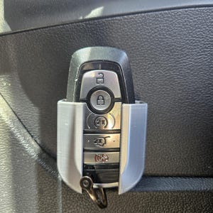 May include: A silver car key with a black plastic case and a silver key ring. The key has buttons for lock, unlock, trunk, and panic. The key is in a holder attached to the inside of a car door.