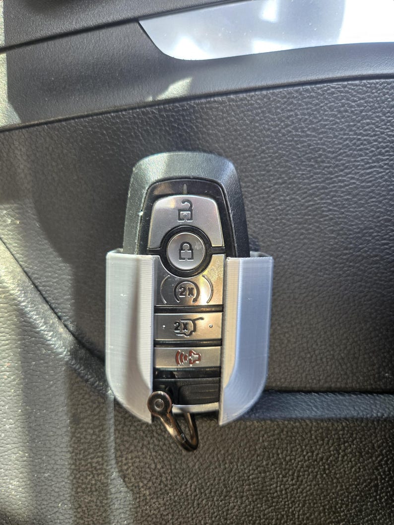 May include: A silver car key with a black plastic case and a silver key ring. The key has buttons for lock, unlock, trunk, and panic. The key is in a holder attached to the inside of a car door.