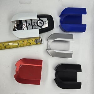 May include: Four key fob cases in different colors: white, silver, red, and black. The cases are designed to protect a car key fob. A tape measure is included for size reference.