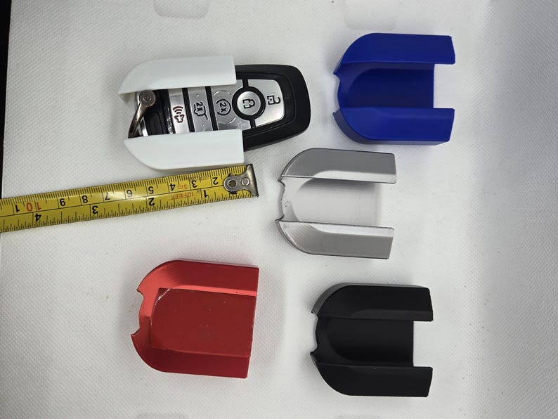 May include: Four key fob cases in different colors: white, silver, red, and black. The cases are designed to protect a car key fob. A tape measure is included for size reference.