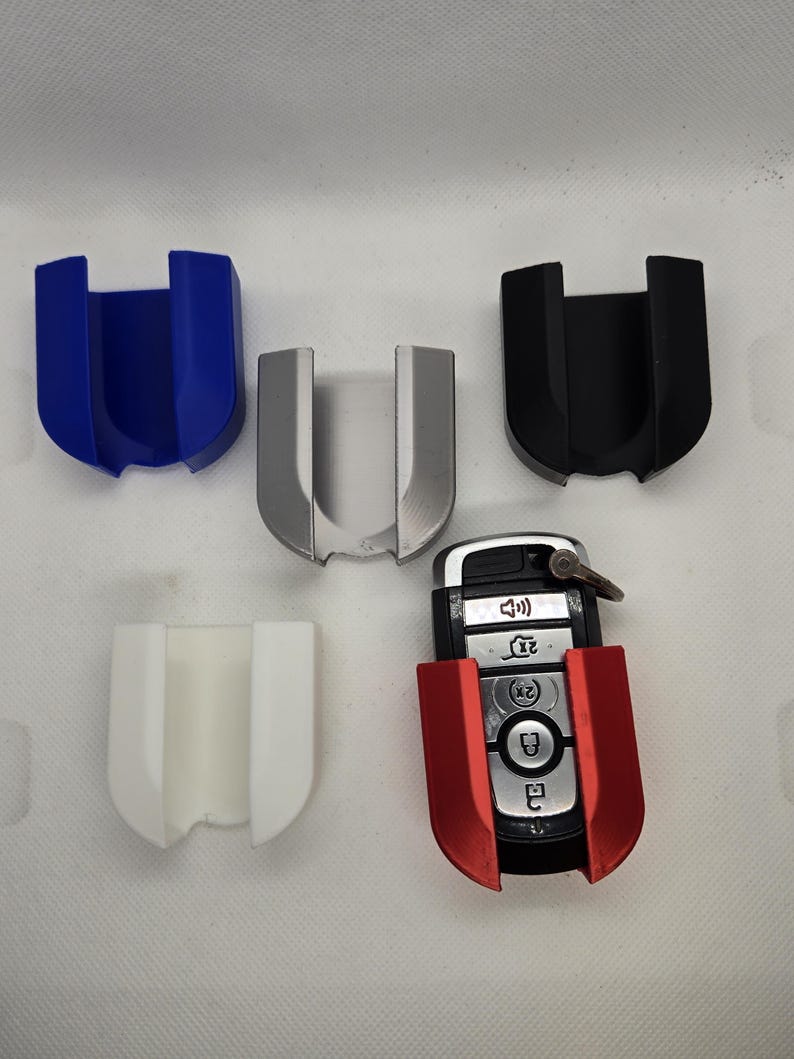 May include: Five key fob holders in different colors: blue, silver, black, white, and red. The red holder is holding a car key fob with buttons.