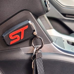 May include: A black and red key chain with the letters "ST" in red. The keychain is attached to a black carbon fiber key fob.