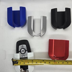 May include: Five key holders in different colors: blue, silver, black, red, and white. The white key holder has a car key inside. A tape measure is placed next to the white key holder.