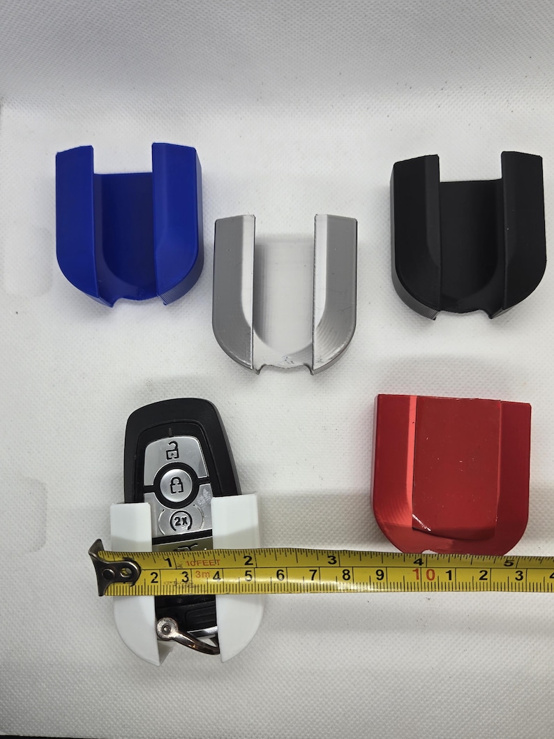 May include: Five key holders in different colors: blue, silver, black, red, and white. The white key holder has a car key inside. A tape measure is placed next to the white key holder.