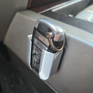 May include: A gray plastic car key holder with a silver car key inside. The holder is attached to the side of a car's center console.
