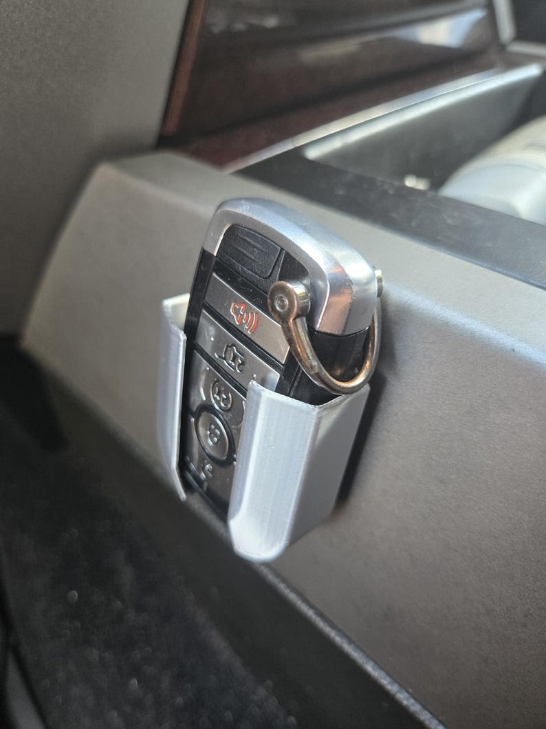 May include: A gray plastic car key holder with a silver car key inside. The holder is attached to the side of a car's center console.