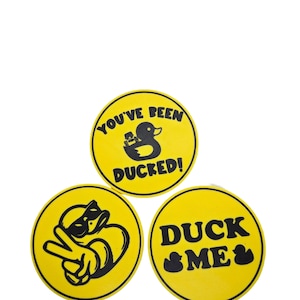 May include: Three yellow circular stickers with black text and graphics. One says "YOU'VE BEEN DUCKED!" with a duck graphic. Another says "DUCK ME" with a duck graphic. The third shows a duck making a peace sign.
