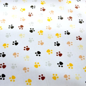 Könnte beinhalten: Weißer Stoff mit einem sich wiederholenden Muster aus Pfotenabdrücken in verschiedenen Farben, darunter Schwarz, Braun, Gelb und Creme.