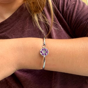 May include: A silver bracelet with a decorative charm. The charm has a purple and white starburst design within a circular frame. The bracelet is worn on a person's wrist.