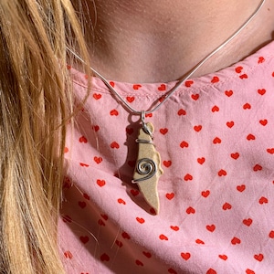 Może przedstawiać: Naszyjnik z łańcuszkiem ze srebra. Wisiorek jest w jasnobrązowym kolorze, ze spiralnym wzorem z drutu. Tło to różowa koszula z czerwonymi sercami.