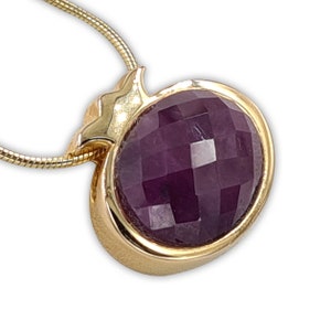 Puede incluir: Collar dorado con colgante. El colgante presenta una gema facetada, de forma ovalada, de color púrpura intenso, engastada en un bisel dorado. Se aprecia un elemento decorativo en forma de hoja. La cadena es una fina cadena de serpiente dorada.