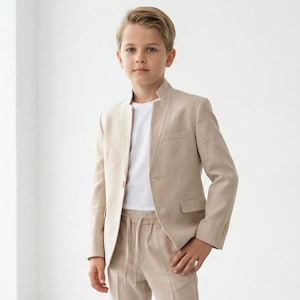 May include: A young person wearing a beige suit with a white t-shirt and white sneakers. The suit includes a blazer and drawstring pants. The outfit is a neutral color and appears to be made of a light fabric.