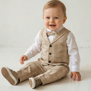 May include: A baby wearing a tan plaid vest, pants, and bow tie, paired with a white button-down shirt. The outfit is completed with matching sneakers. The baby is smiling and sitting on a white surface.