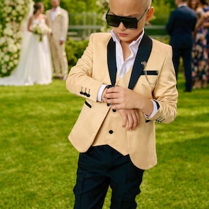 May include: A young person in a tan suit with black lapels, a white shirt, and black pants, wearing sunglasses. The suit has a matching vest and is accessorized with a chain. The background shows a wedding ceremony on a green lawn.