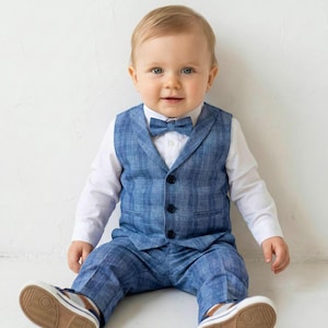 May include: A baby wearing a blue plaid vest, matching pants, and a bow tie over a white dress shirt. The outfit is paired with white and brown sneakers. The baby is sitting and smiling.