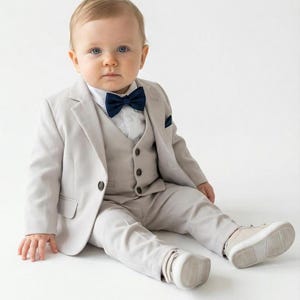 May include: A baby wearing a light gray suit, vest, and pants, paired with a navy blue bow tie and matching pocket square. The outfit is completed with white sneakers. The baby is sitting on a white surface.