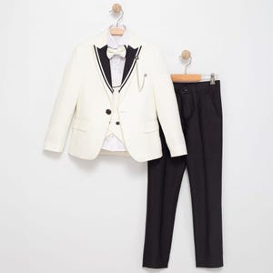 May include: A formal ivory suit for a child, comprising a blazer, waistcoat, dress shirt, bow tie, and black trousers. The blazer has black trim and a decorative chain. The suit is hung on a white wall.