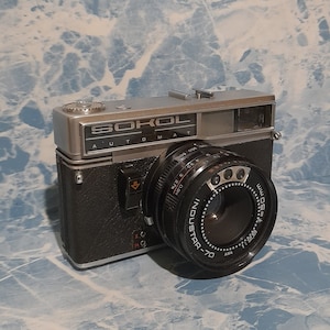 Puede incluir: Una cámara de película de 35 mm de color plateado y negro con el texto "Sokol Automat" en la parte superior. La cámara tiene una lente negra con el texto "Industar-70" en ella.