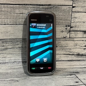 May include: A silver and black Nokia smartphone with a screen showing a blue and teal geometric design. The phone's screen displays the word "Автономный" and the date 03.02.2011. The phone is on a wooden surface.