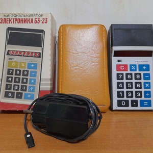 Peut inclure: Calculatrice Elektronika B3-23 vintage avec boîte et étui d'origine. La calculatrice a un clavier noir et blanc avec des touches de fonction rouges et bleues. La boîte porte le texte "МИКРОКАЛЬКУЛЯТОР ЭЛЕКТРОНИКА БЗ-23". Un cordon d'alimentation noir est également inclus.