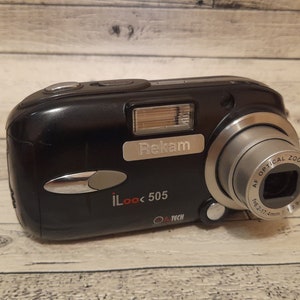 May include: A black digital camera with a silver lens and a silver button on the front. The camera has the text "iLook 505" and "Rekam" on the front.