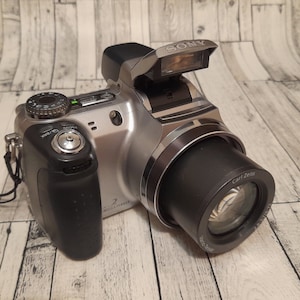 May include: A silver Sony digital camera with a black Carl Zeiss lens. The camera has a built-in flash and a black hand grip.