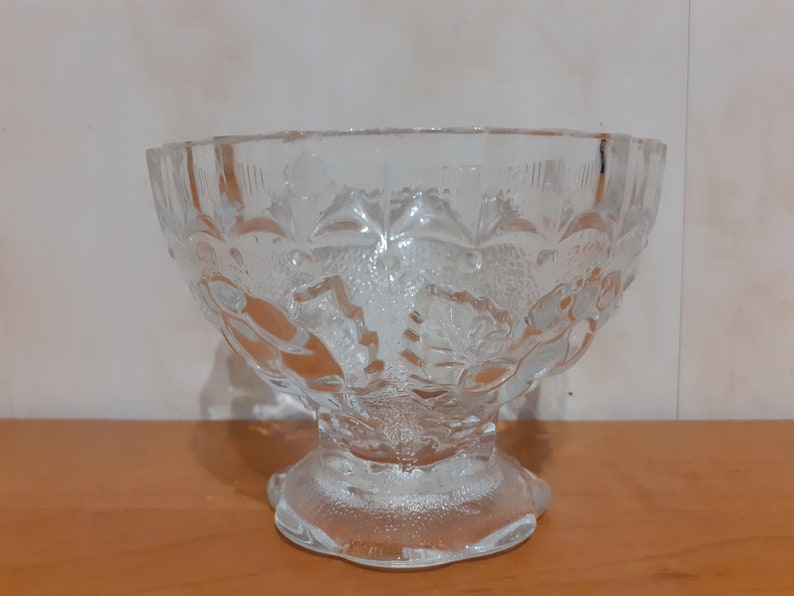 May include: A clear glass bowl with a decorative floral pattern. The bowl has a wide, round base and a flared rim.