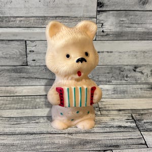 Op de afbeelding: Een vintage, lichtbeige berenbeeldje met een kleurrijk accordeon. De beer heeft zwarte ogen en een zwarte neus, met een rode mond. Het accordeon heeft rode, groene en witte details. De beer draagt een luier met blauwe en groene stippen.