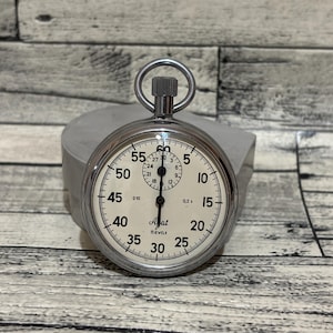 May include: A silver pocket watch with a white face and black markings. The watch face has numbers from 5 to 60, and the words "Ayat" and "Bewels" are printed on the face.
