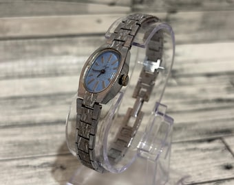 May include: A vintage silver-toned wristwatch with a light blue oval face. The watch has a textured metal band and a small, dark-colored crown. The watch is displayed on a clear plastic stand.