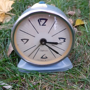 Pode incluir: Um despertador vintage azul com um mostrador branco e ponteiros pretos. O relógio tem uma moldura cromada e um botão branco no topo. O mostrador do relógio diz "Сальск СССР" e "Сальск".