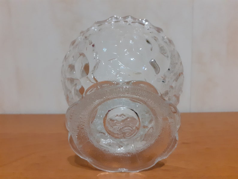 May include: A clear glass bowl with a textured design and a pedestal base. The bowl is empty and appears to be made of thick glass.