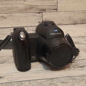May include: A black Konica Minolta digital camera with a lens cap on. The camera has a strap attached and a small black accessory attached to the side.