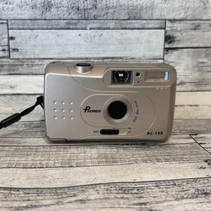 Może przedstawiać: Srebrny aparat fotograficzny Premier PC-135 na film z czarnym obiektywem i lampą błyskową. Aparat posiada czarny pasek na nadgarstek, a na przodzie widnieją napisy "Premier" i "PC-135". Obiektyw to obiektyw 30 mm.