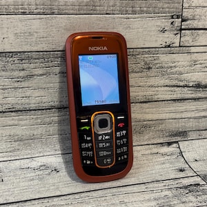 May include: A red Nokia mobile phone with a black keypad and a rectangular screen displaying the time 09:00 and the date 01.01.2008. The device features a central navigation button surrounded by an orange border and standard alphanumeric keys.