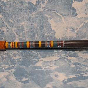 Op de afbeelding: Een houten rookpijp met een bruine kom en een lange, gebogen steel. De steel is versierd met een kleurrijk gestreept patroon in tinten blauw, geel, rood en zwart.