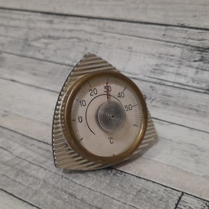 Op de afbeelding: Een zilveren en gouden analoge thermometer met een wit gezicht en zwarte markeringen. De thermometer geeft aan van 0 tot 50 graden Celsius. De naald wijst naar 30 graden.