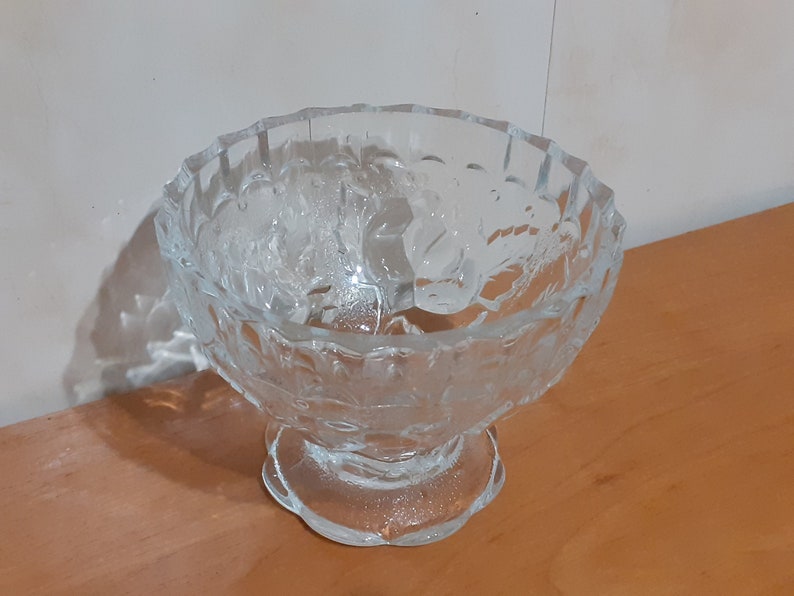 May include: A clear glass bowl with a pedestal base. The bowl has a decorative, embossed pattern of leaves and flowers.