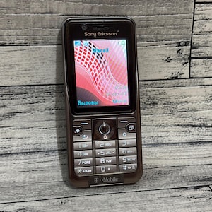 Op de afbeelding: Een bruine Sony Ericsson mobiele telefoon met een toetsenbord en een scherm met een roze en rode afbeelding. De telefoon heeft het T-Mobile-logo en het woord "Вызовы" op het scherm.