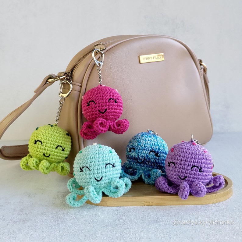May include: Five crocheted octopus keychains in different colours, including pink, green, blue, and purple, hanging from a brown leather handbag. The octopus keychains have black eyes and smiling mouths.