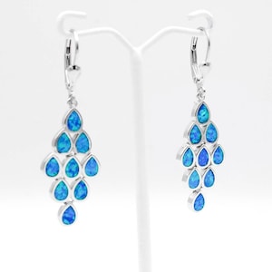 May include: Silver chandelier earrings with blue teardrop-shaped opals. Each earring features a cascade of opal stones, creating a shimmering effect. The earrings are displayed on a white stand against a white background.