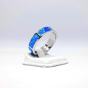 Puede incluir: Un anillo de banda de plata con una incrustación de ópalo azul. El anillo se muestra en un soporte de anillo blanco.