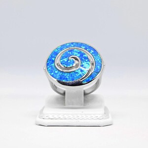 Op de afbeelding: Een zilveren ring met een ronde, blauwe opaalsteen. De opaal heeft een spiraalvormig ontwerp in een lichtere blauwtint. De ring wordt gepresenteerd op een witte standaard tegen een witte achtergrond. De ring is een sieraad.