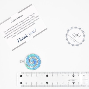 K&ouml;nnte beinhalten: Ein runder silberner Anh&auml;nger mit einer leuchtend blauen und gr&uuml;nen irisierenden Einlage in Spiralform, platziert &uuml;ber einem transparenten Lineal, das einen Durchmesser von etwa 3,2 cm zeigt, neben einer Dankeskarte und einem Logo.