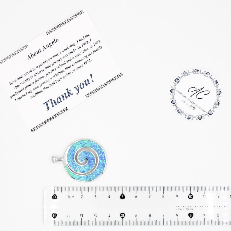 K&ouml;nnte beinhalten: Ein runder silberner Anh&auml;nger mit einer leuchtend blauen und gr&uuml;nen irisierenden Einlage in Spiralform, platziert &uuml;ber einem transparenten Lineal, das einen Durchmesser von etwa 3,2 cm zeigt, neben einer Dankeskarte und einem Logo.
