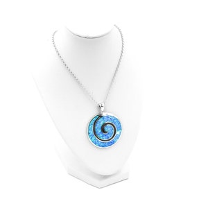 K&ouml;nnte beinhalten: Eine silberfarbene Halskette mit einem runden Anh&auml;nger, der eine leuchtend blaue, irisierende Einlage aufweist. Der Anh&auml;nger zeigt ein markantes schwarzes Spiraldesign in der Mitte des blauen Materials und h&auml;ngt an einer passenden Gliederkette.