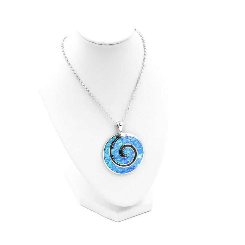 K&ouml;nnte beinhalten: Eine silberfarbene Halskette mit einem runden Anh&auml;nger, der eine leuchtend blaue, irisierende Einlage aufweist. Der Anh&auml;nger zeigt ein markantes schwarzes Spiraldesign in der Mitte des blauen Materials und h&auml;ngt an einer passenden Gliederkette.