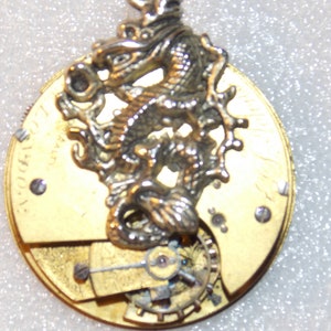 May include: A gold-toned pocket watch with a silver dragon charm attached to the top. The watch face is visible and has a gold-toned background with black markings. The watch is open and the inner workings are visible.