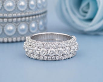 May include: A silver-toned band ring featuring three rows of clear, round-cut stones. The center row consists of larger stones, while the top and bottom rows are composed of smaller, pavé-set stones that create a continuous, sparkling appearance.