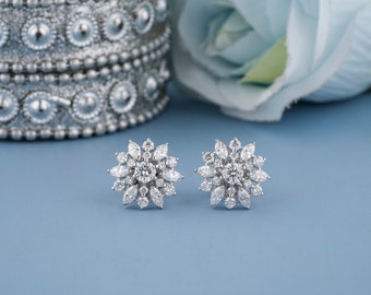 May include: A pair of sparkling, silver-toned stud earrings. Each earring features a central, round, clear gemstone surrounded by smaller, marquise-cut stones, creating a floral or starburst design. The earrings are set against a light blue backdrop.